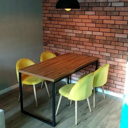 Wykończenia Marcin Kieryk Działalność usługowa - Jadalnia z drewnianym stołem na czarnych nogach i czterema żółtymi krzesłami na tle ściany z cegły, oświetlona czarną lampą wiszącą.