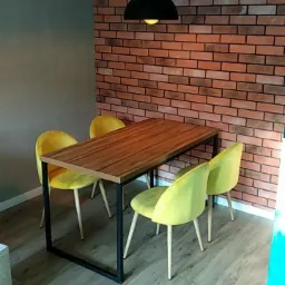 Jadalnia z drewnianym stołem na czarnych nogach i czterema żółtymi krzesłami na tle ściany z cegły, oświetlona czarną lampą wiszącą.