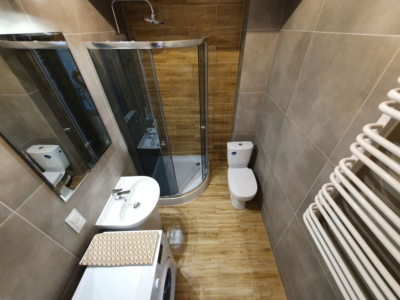 Nowoczesna łazienka z kabiną prysznicową, umywalką nablatową, toaletą i grzejnikiem drabinkowym, wykończona płytkami w odcieniach brązu i beżu, z drewnopodobną podłogą i pralką pod umywalką...