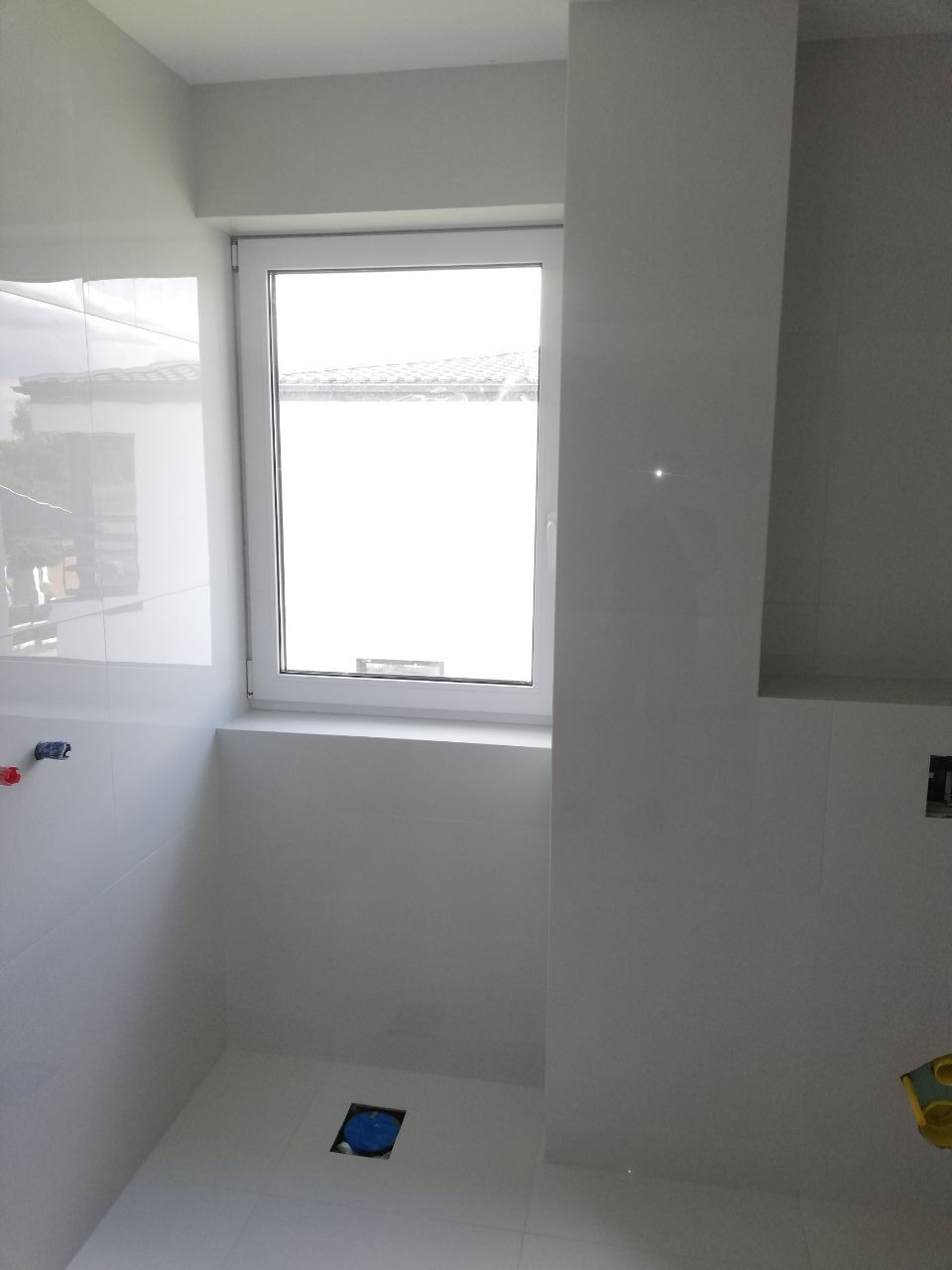 Wnętrze łazienki w trakcie prac wykończeniowych, widoczne białe płytki na ścianach i podłodze, okno z widokiem na zewnątrz oraz otwór odpływowy w podłodze zabezpieczony niebieską folią.