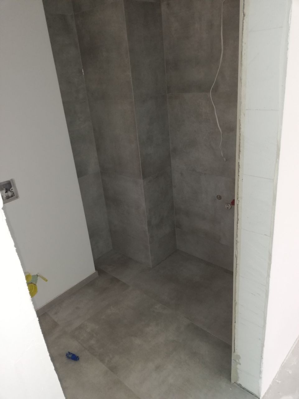 Wnętrze łazienki w trakcie prac wykończeniowych, widoczne szare płytki na ścianach i podłodze, surowa ściana z instalacją, fragment narzędzia na podłodze.
