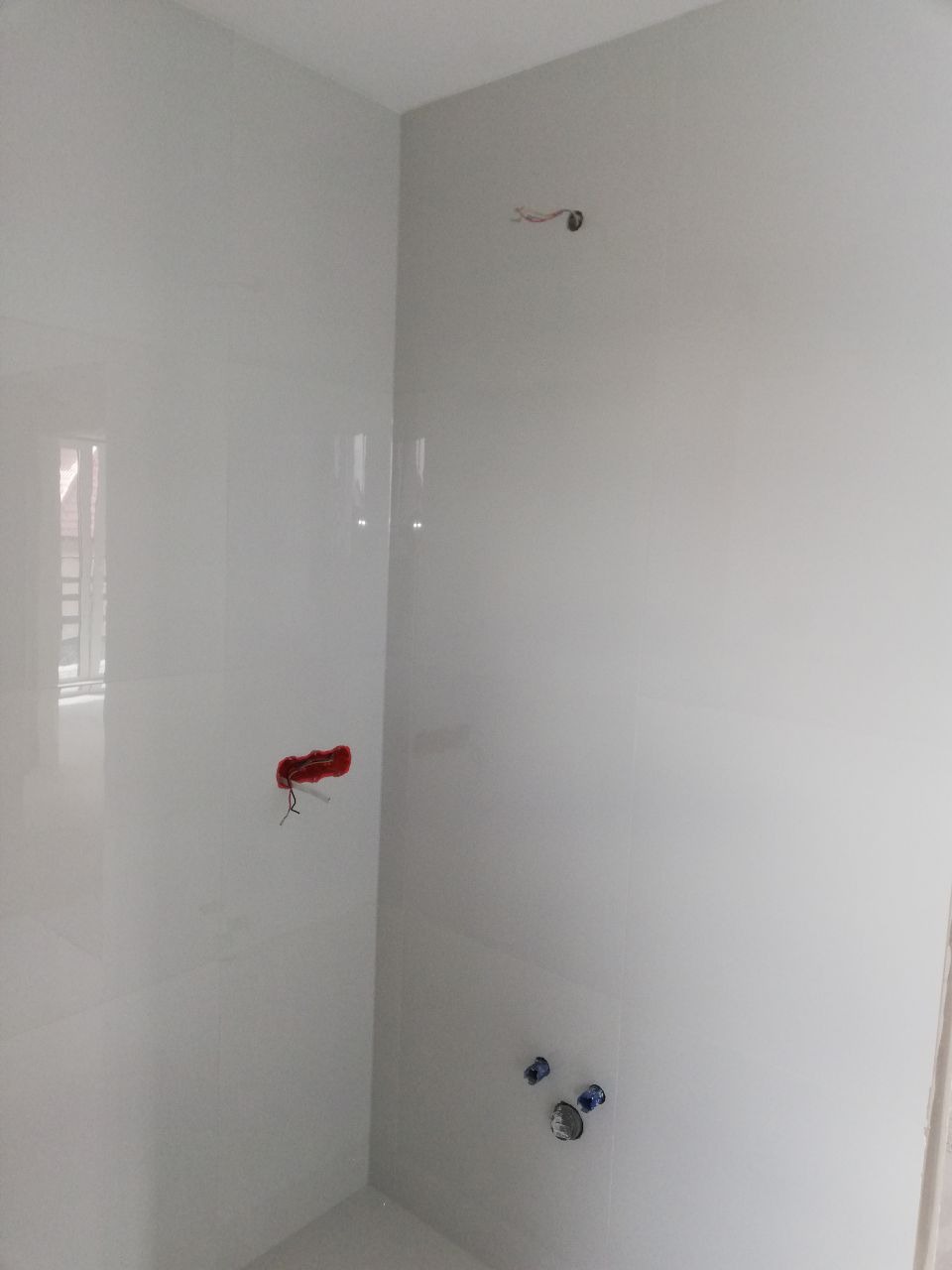 Ściana łazienki wyłożona błyszczącymi, białymi płytkami, widoczne wystające przewody elektryczne, puszka instalacyjna w kolorze czerwonym oraz przyłącza wodne.