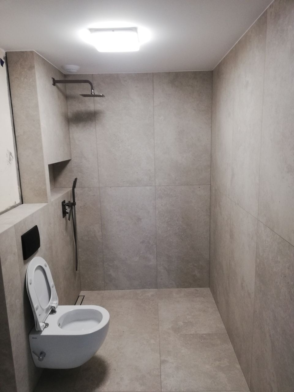 Nowoczesna łazienka z beżowymi, matowymi płytkami imitującymi kamień, wisząca toaleta z otwartą klapą, deszczownica i półka wnękowa, oświetlenie sufitowe.