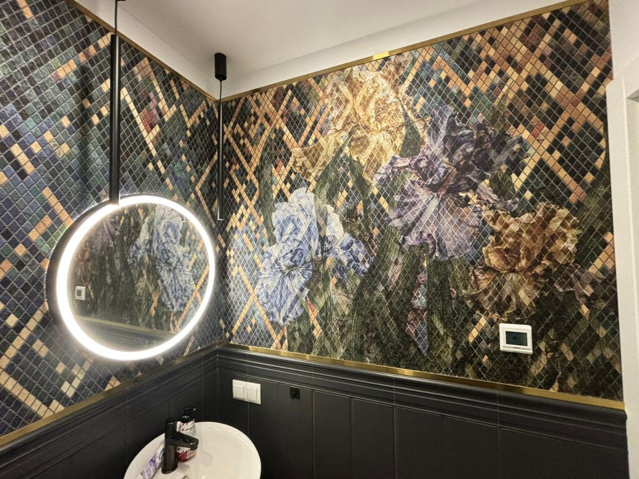 Elegancka łazienka z mozaiką w kwiatowe wzory, okrągłe lustro z podświetleniem LED, czarne płytki do połowy ściany z złotym akcentem, biała umywalka.