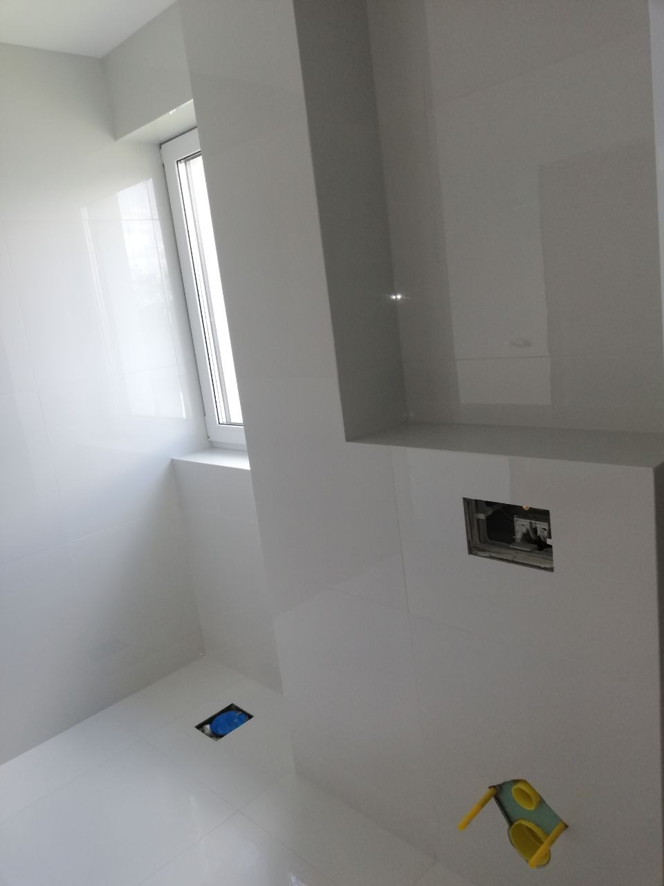 Białe, błyszczące płytki w łazience, widoczne otwory na instalacje, fragment okna z parapetem.