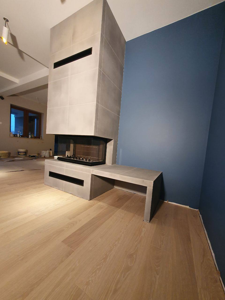 Nowoczesny kominek obłożony szarymi płytami, z czarnym wkładem kominkowym i przylegającą ławką, na tle pomalowanej na granatowo ściany i parkietu w jasnym odcieniu drewna.