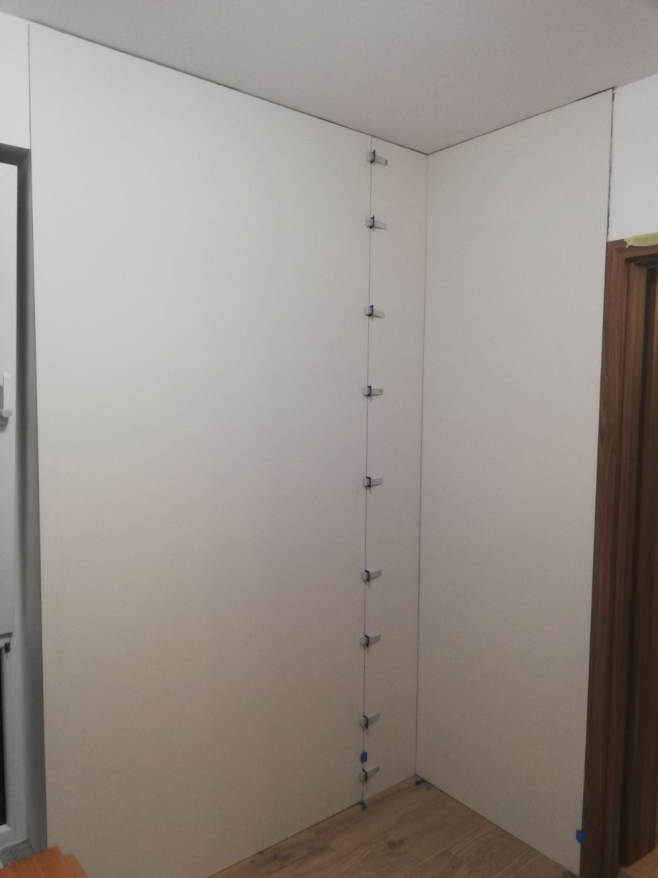Biała ściana z zamontowanymi płytami gipsowo-kartonowymi, pionowy rząd metalowych klipsów utrzymujących płyty na miejscu, widoczne łączenia płyt, podłoga z paneli.