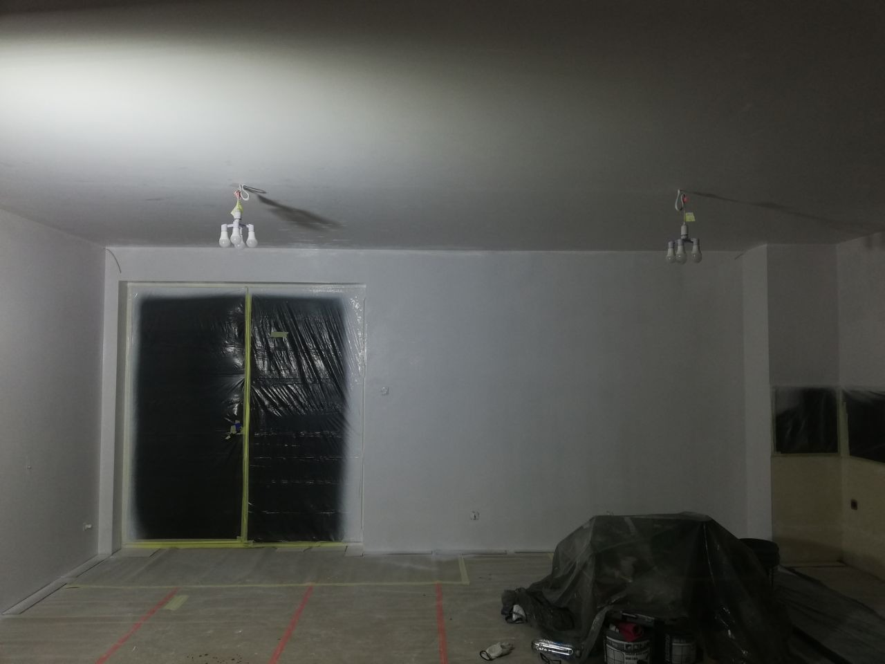 Pokój w trakcie malowania: ściany pomalowane na szaro, podłoga zabezpieczona folią, drzwi osłonięte czarną folią malarską, widoczne puszki z farbą i prowizoryczne oświetlenie.