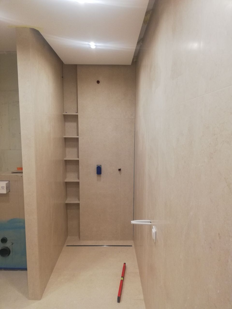 Wnętrze łazienki w trakcie wykańczania, widoczne beżowe płytki, wnęka z półkami, odpływ liniowy w podłodze oraz poziomica na podłodze, sugerująca prace glazurnicze.