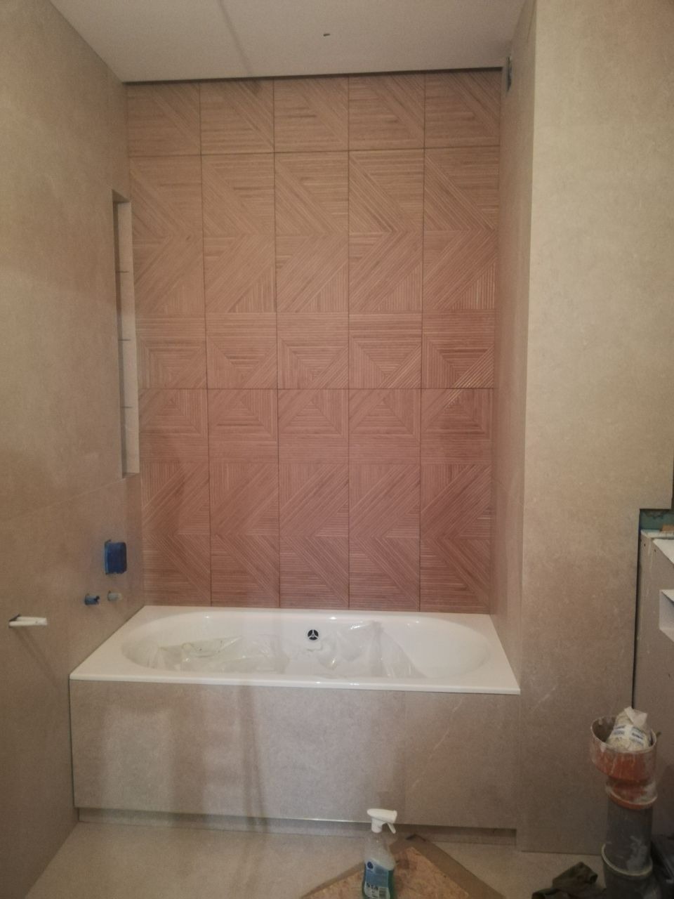 Ściana nad wanną wyłożona kwadratowymi płytkami z geometrycznym wzorem w odcieniach beżu i brązu, wanna obudowana płytkami w kolorze ściany, widoczne akcesoria i chemia gospodarcza na podłodze.