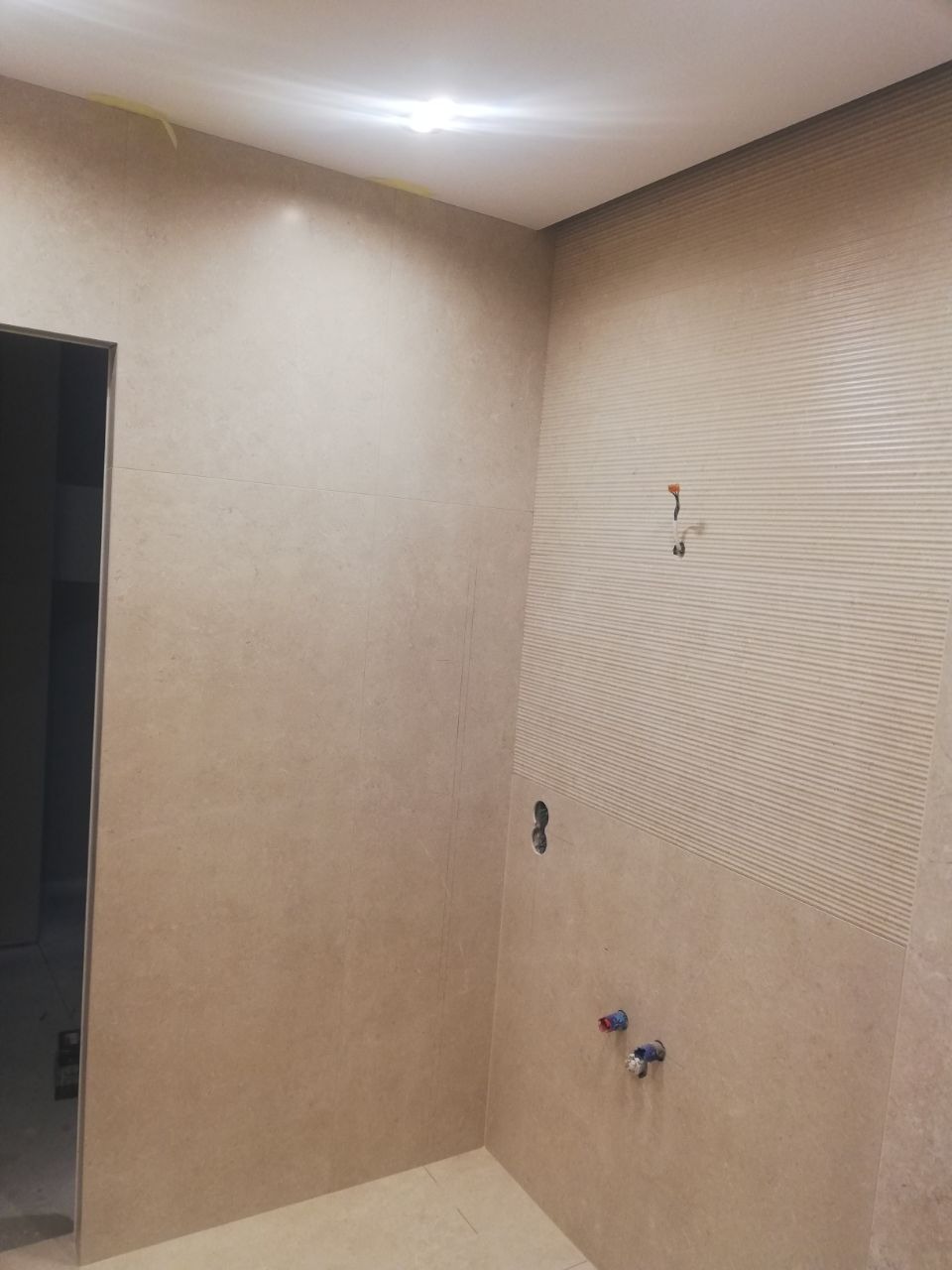 Układanie beżowych płytek w łazience w Łańcucie, widoczne przygotowane otwory na armaturę i pojedynczy przewód elektryczny wystający ze ściany, część ściany z poziomymi żłobieniami.