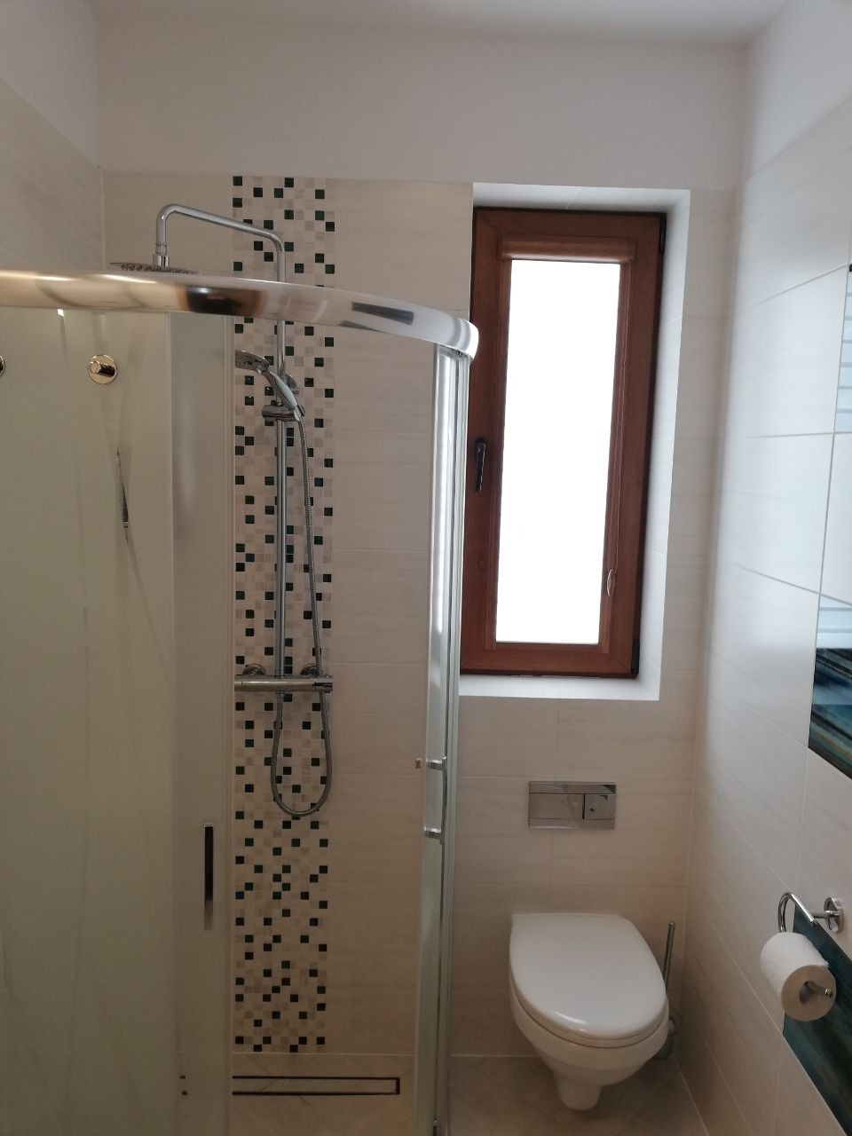 Jasna łazienka z kabiną prysznicową typu walk-in, mozaiką z czarnych kwadratów przy prysznicu, drewnianą ramą okna i podwieszaną toaletą.