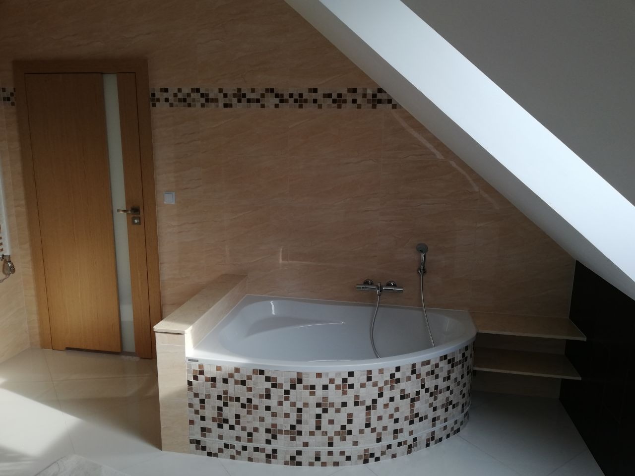 Nowoczesna łazienka na poddaszu ze skośnym sufitem, beżowe płytki na ścianach z dekoracyjnym pasem mozaiki, narożna wanna obudowana mozaiką z brązowych i beżowych kwadratów, jasne drewniane drzwi...