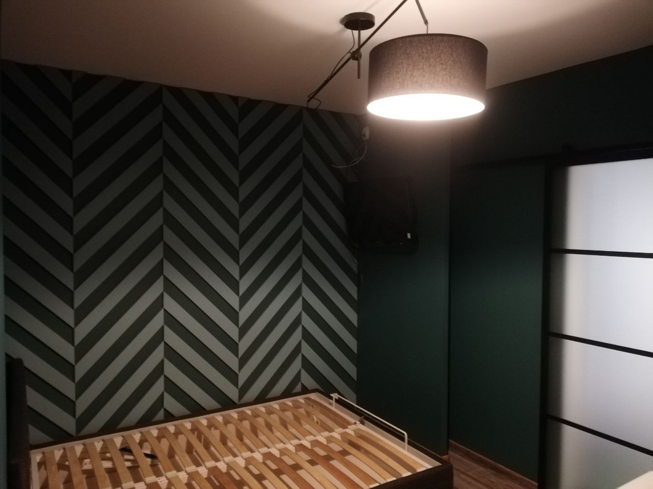 Pokój z dekoracyjną ścianą w jodełkę w odcieniach szarości i zieleni, widoczny stelaż łóżka bez materaca, oświetlony lampą sufitową z abażurem w kolorze szarym.