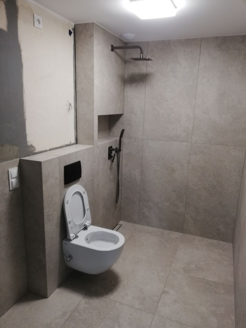 Wnętrze łazienki w trakcie remontu: zawieszona toaleta z otwartą klapą, szara płytka na ścianach i podłodze, widoczna instalacja prysznicowa i fragment ściany przed wykończeniem.