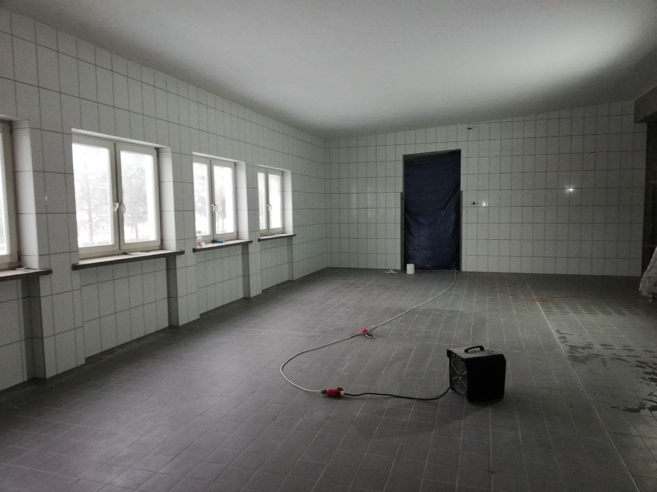 Duże, puste pomieszczenie wyłożone szarymi płytkami podłogowymi i białymi płytkami ściennymi, z trzema oknami po lewej stronie, zakrytymi parapetami, zasłoniętymi drzwiami z tyłu i grzejnikiem...