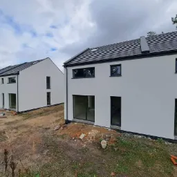 Dwa nowo wybudowane, bliźniacze domy z jasną elewacją i grafitową stolarką okienną na nierównym terenie przed zagospodarowaniem.