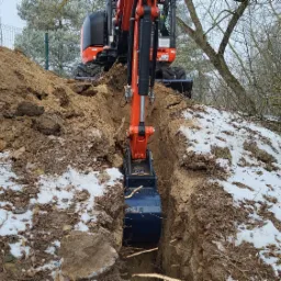 Pomarańczowa mini koparka w Tomaszowie Mazowieckim kopie wąski wykop w ziemi z resztkami śniegu, widok z perspektywy operatora, na drugim planie sterta ziemi i ogrodzenie z siatki.