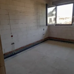 Surowy stan wnętrza z widocznymi przewodami elektrycznymi w ścianach z betonu komórkowego i rurami instalacyjnymi przy podłodze, okno otwarte na zewnątrz budynku.