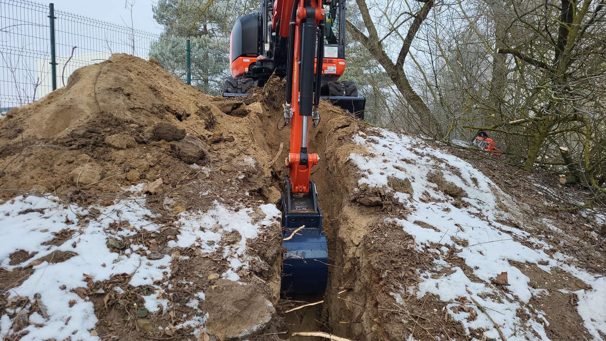 Pomarańczowa mini koparka w Tomaszowie Mazowieckim kopie wąski wykop w ziemi z resztkami śniegu, widok z perspektywy operatora, na drugim planie sterta ziemi i ogrodzenie z siatki.