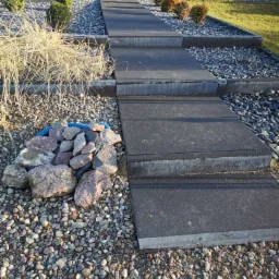 Nowoczesne schody zewnętrzne z szarych płyt betonowych prowadzące do domu, otoczone ozdobnym żwirem i niskimi krzewami.