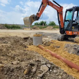 Pomarańczowa koparka Hitachi Zaxis 26U zasypuje piaskiem rurę drenarską w wykopie, widoczny betonowy krąg studzienny wypełniony piaskiem, w tle droga i błękitne niebo z chmurami.