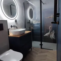 Nowoczesna łazienka z owalnym lustrem z podświetleniem LED, umywalką nablatową na szafce z drewnianym blatem, kabiną prysznicową z motywem czapli i podłogą ułożoną w jodełkę.