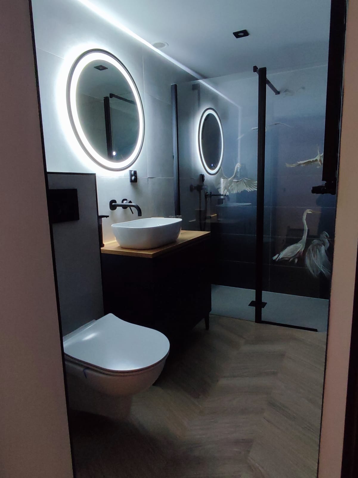 Nowoczesna łazienka z podświetlanymi okrągłymi lustrami, czarną armaturą, umywalką nablatową na drewnianym blacie, toaletą wiszącą i kabiną prysznicową z motywem czapli na czarnych płytkach.
