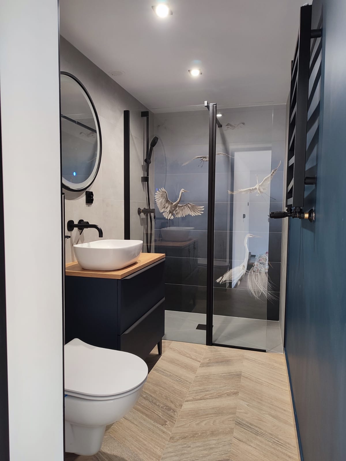 Nowoczesna łazienka z kabiną prysznicową zdobioną motywem ptaków, umywalką nablatową na szafce w kolorze granatowym, okrągłym lustrem z podświetleniem LED i grzejnikiem drabinkowym w kolorze...