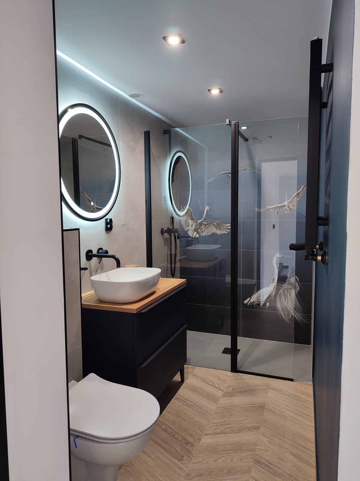 Nowoczesna łazienka z okrągłymi lustrami podświetlanymi LED, umywalką nablatową na czarnej szafce z drewnianym blatem, kabiną prysznicową z motywem żurawi i parkietem ułożonym w jodełkę.