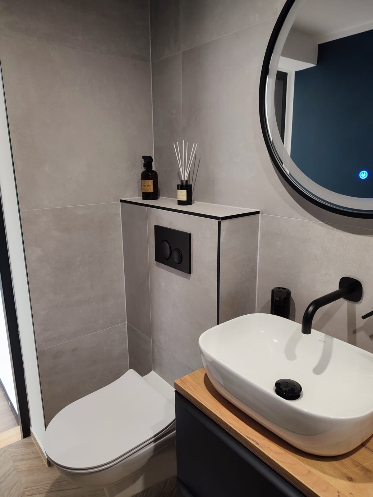 Nowoczesna łazienka z szarymi płytkami, podwieszaną toaletą, umywalką nablatową na drewnianym blacie i okrągłym lustrem z podświetleniem LED.