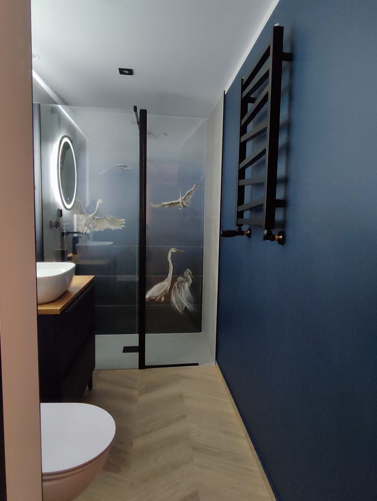 Nowoczesna łazienka z umywalką nablatową, podświetlanym lustrem i kabiną prysznicową z motywem ptaków, ciemnoniebieska ściana z czarnym grzejnikiem drabinkowym, parkiet ułożony w jodełkę.