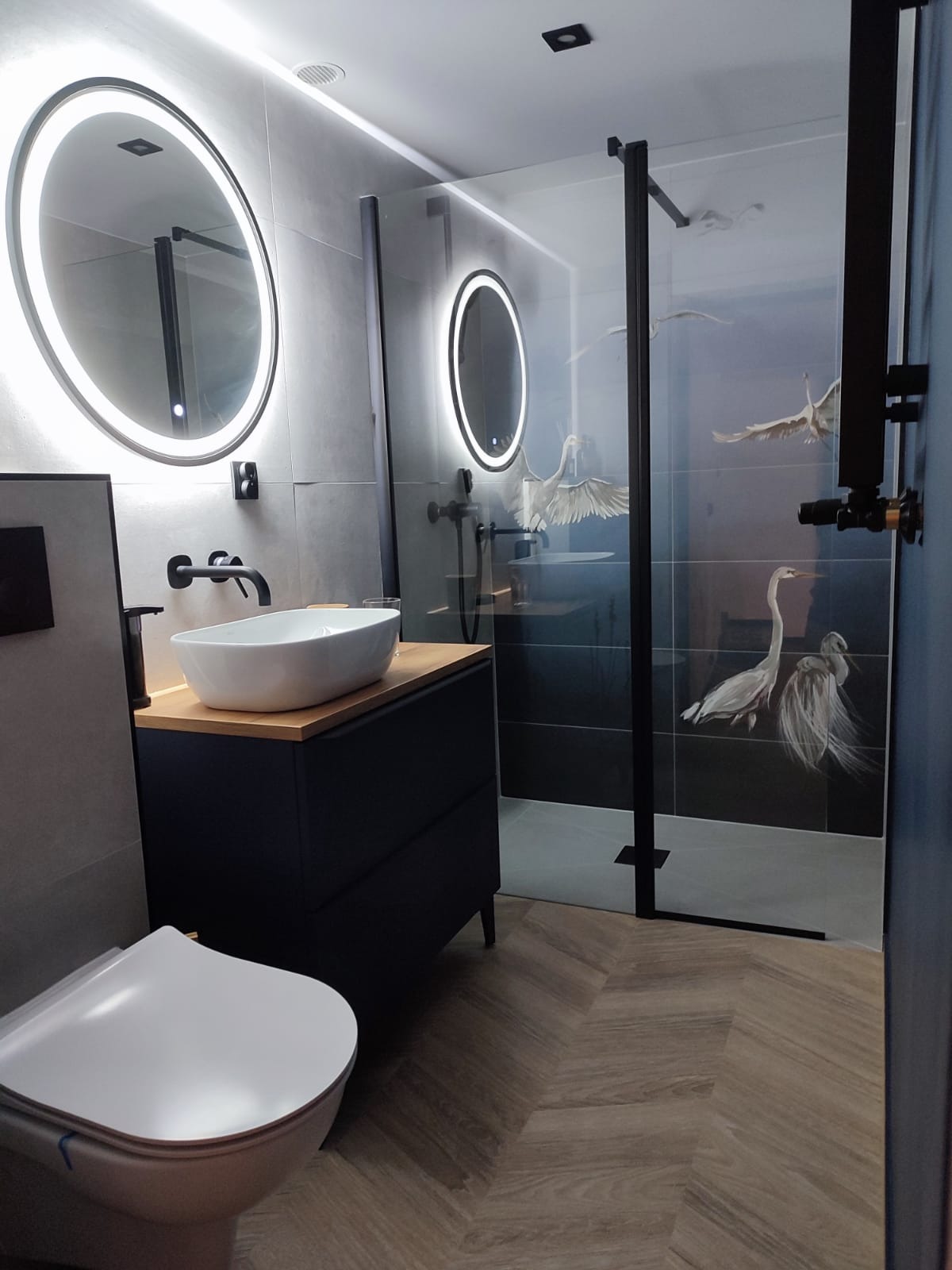 Nowoczesna łazienka z owalnym lustrem z podświetleniem LED, umywalką nablatową na szafce z drewnianym blatem, kabiną prysznicową z motywem czapli i podłogą ułożoną w jodełkę.