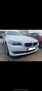 Białe BMW serii 5 na brukowanym parkingu, widoczne światła przednie i grill, zarys czerwonego samochodu w tle.