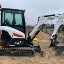 Mini koparka Bobcat E27 na placu budowy, z widoczną łyżką, gąsienicami i kabiną operatora, obok pryzma piasku i ogrodzenie z siatki.