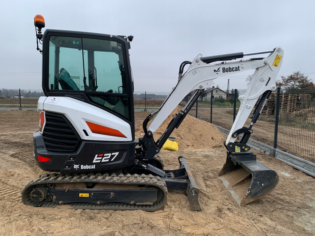 Mini koparka Bobcat E27 na placu budowy, z widoczną łyżką, gąsienicami i kabiną operatora, obok pryzma piasku i ogrodzenie z siatki.