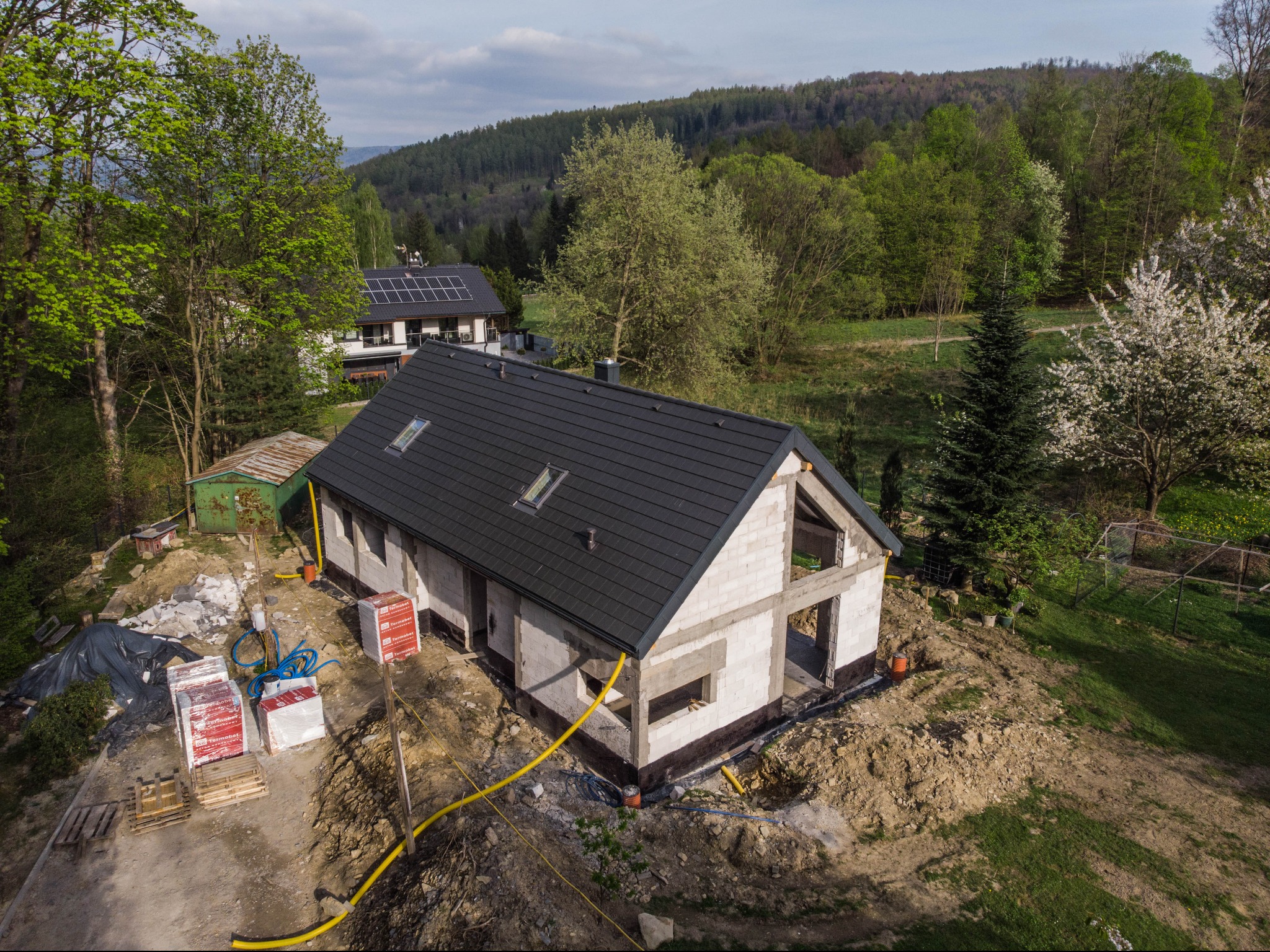 Widok z drona na surowy, dwukondygnacyjny dom z białego betonu komórkowego pod czarną dachówką, w trakcie budowy, z widocznymi paletami bloczków Termobet i żółtymi rurami gazowymi, na tle zieleni...