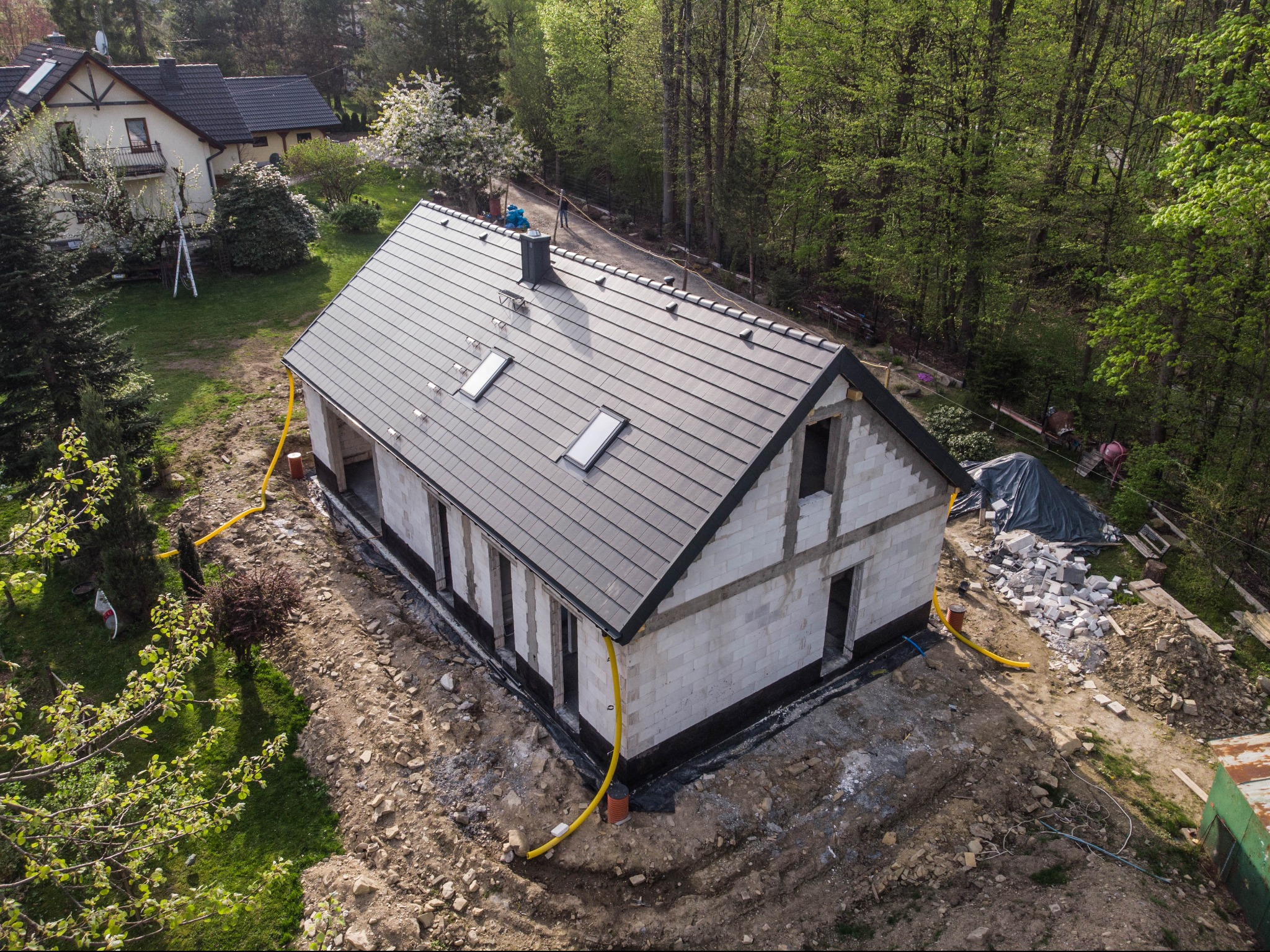 Surowy stan domu z bloczków, z szarą dachówką i oknami dachowymi, widok z góry na plac budowy, z żółtymi rurami odprowadzającymi wodę i stertą materiałów budowlanych w tle.