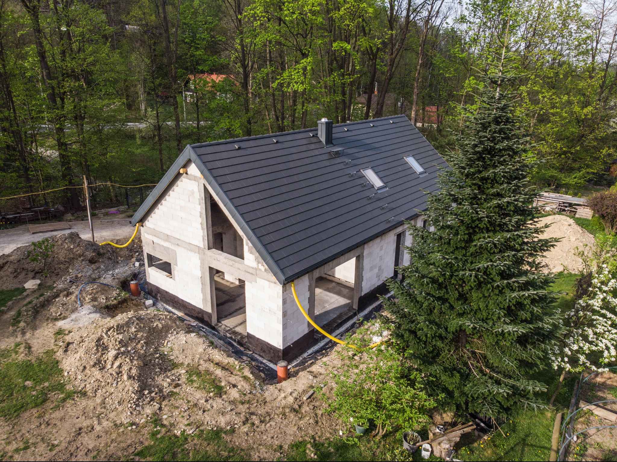 Surowy stan domu z pustaków, z zamontowaną ciemną dachówką, widok z góry na tle zieleni, żółte rury przy ścianach.