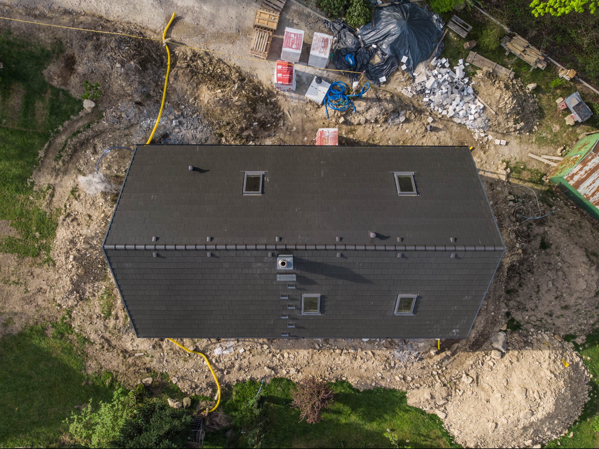 Widok z góry na dach domu z ciemnej dachówki z oknami dachowymi, teren wokół w trakcie prac ziemnych, widoczne materiały budowlane, palety i węże.