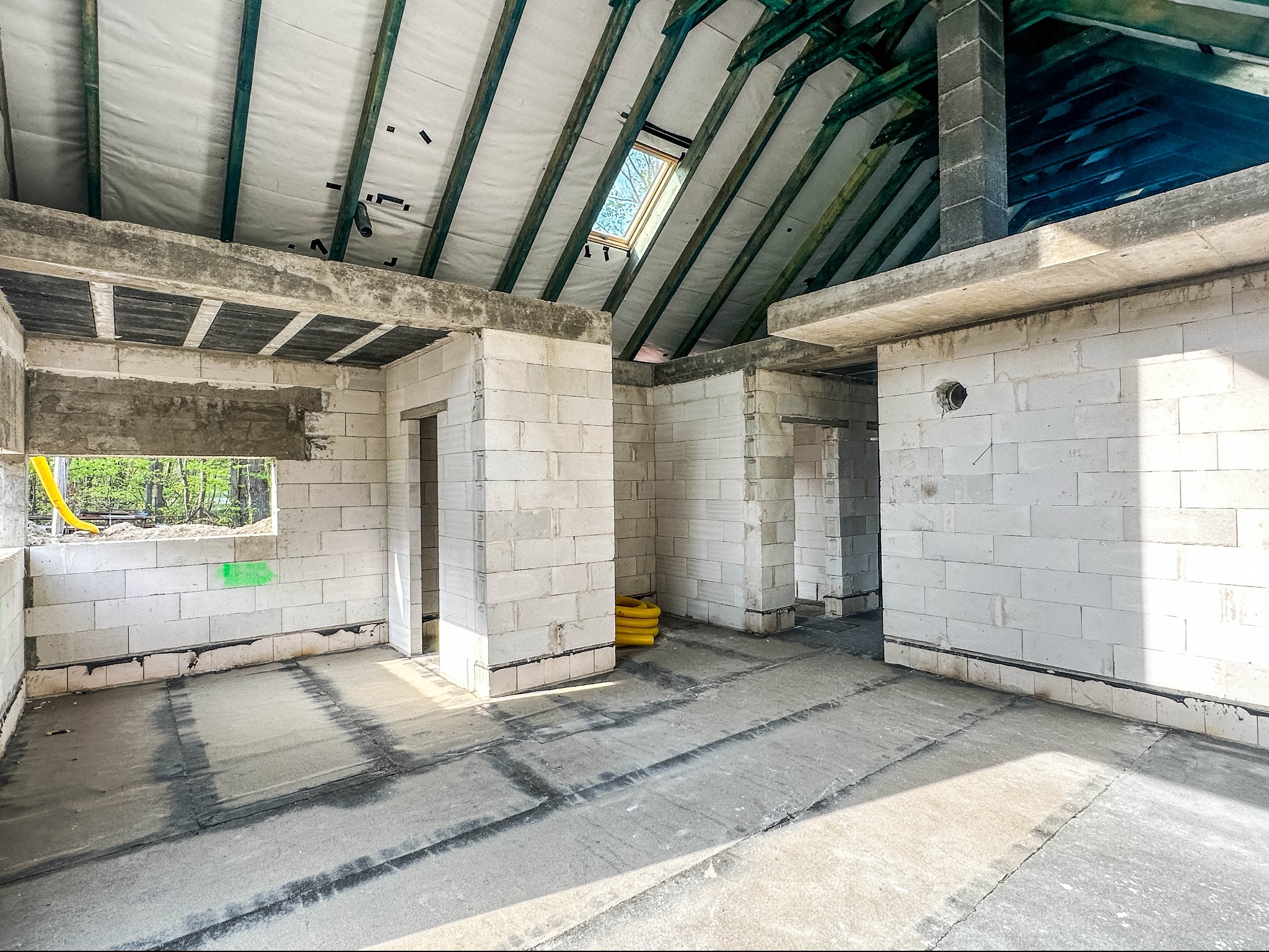 Wnętrze surowego domu z białych bloczków, widoczne elementy konstrukcji dachu i okno dachowe, żółty wąż na podłodze.