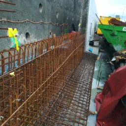 Zbrojenie pionowej ściany oporowej, widok na siatkę z prętów stalowych, betonową ścianę z otworami i zielony kontener na gruz w tle.