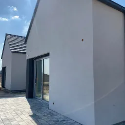 Dwa budynki jednorodzinne z jasną elewacją i grafitową stolarką okienną, połączone brukowanym tarasem z kostki w odcieniach szarości, w tle błękitne niebo z promieniami słońca.