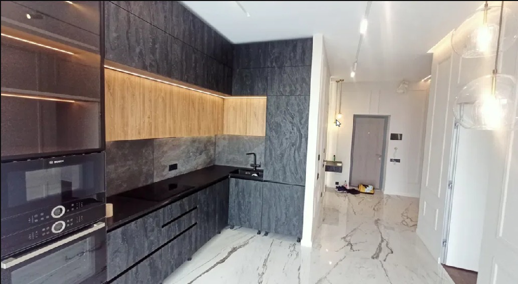 Nowoczesna kuchnia z szarymi i drewnianymi szafkami, czarnym blatem i marmurową podłogą, oświetlona listwami LED pod szafkami wiszącymi i wiszącymi lampami szklanymi w sąsiednim pomieszczeniu.