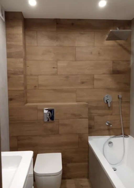 Nowoczesna łazienka z drewnopodobnymi płytkami na ścianach, białą armaturą i minimalistycznym prysznicem deszczownicą.