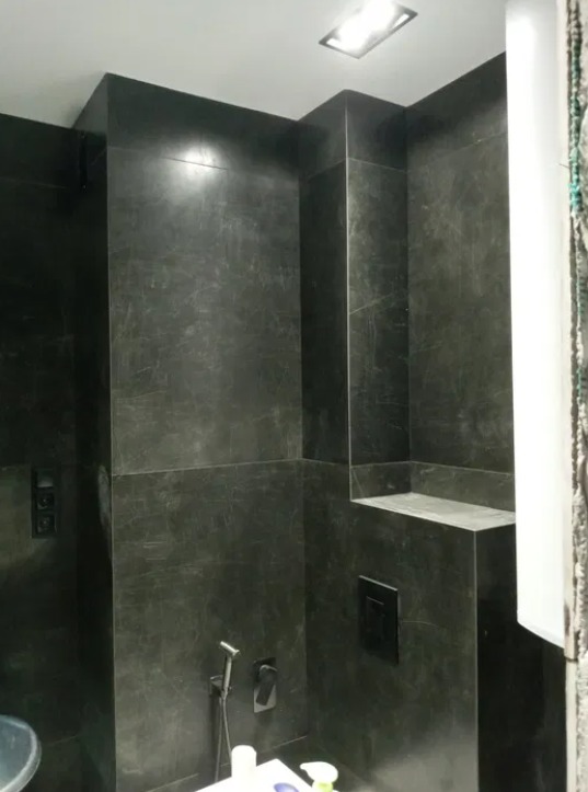 Wykończona łazienka z ciemnymi, marmurkowymi płytkami, widoczna armatura, wnęka w ścianie i podwieszana toaleta.