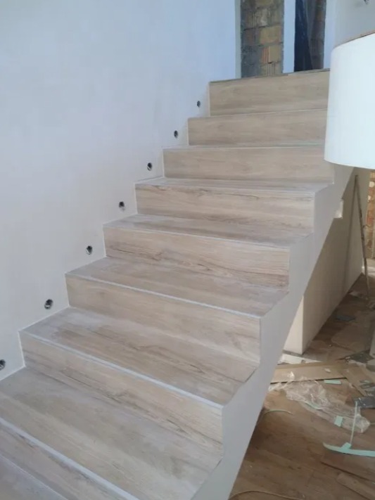 Schody wewnętrzne wykończone panelami imitującymi jasne drewno, z zamontowanymi punktowymi lampkami w ścianie, widoczne przygotowania do dalszych prac wykończeniowych.