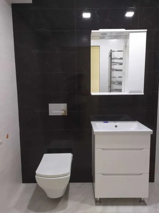 Nowoczesna łazienka z czarnymi płytkami imitującymi marmur, białą ceramiką sanitarną i szafką pod umywalką z trzema szufladami na metalowych nóżkach, widoczne odbicie grzejnika drabinkowego w lustrze.