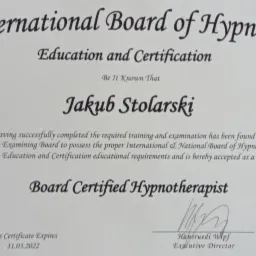 Certyfikat Board Certified Hypnotherapist wydany przez International Board of Hypnosis dla Jakuba Stolarskiego, z datą ważności 31.03.2022, na tle dywanu.