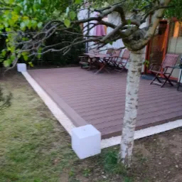 Brązowy taras z desek kompozytowych obramowany białym żwirem i blokami, częściowo zacieniony przez brzozę z widocznym zestawem mebli ogrodowych.